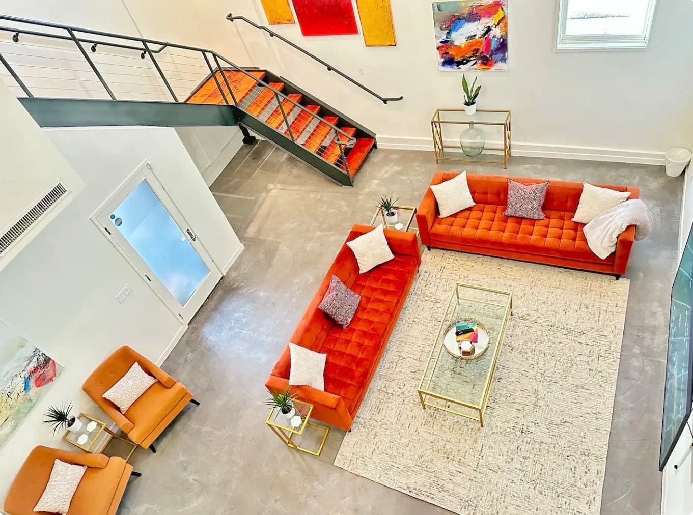 Two-story loft living room with orange velvet sofas, glass coffee table, staircase and colorful wall art